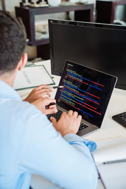 A close-up view of a modern, slim silver laptop on a white desk, displaying a dark-themed code editor with multicolored lines of programming code. The laptop is positioned centrally, with a smartphone