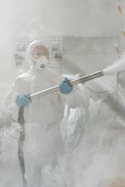 A person dressed in full protective cleaning gear, including a white coverall, face mask, goggles, and blue gloves, is operating a high-powered steam or fogging device emitting a dense cloud of disinf